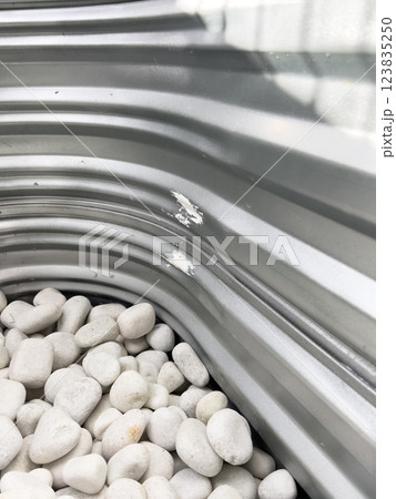 A close-up view of a corrugated metal window well with white decorative rocks at the base. The metal surface has minor scratches and wear, reflecting natural light. This functional design is commonly 123835250