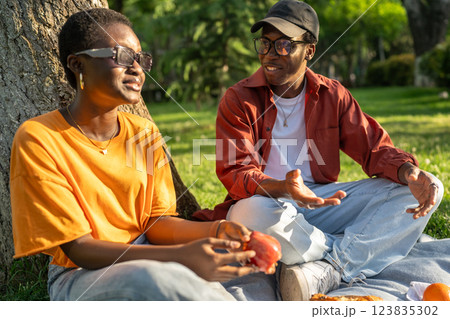 Black couple enjoys peaceful picnic, sit on blanket savoring snacks in conversation, date outdoors Black couple enjoys peaceful picnic, sit on blanket savoring snacks in conversation, date outdoors 123835302