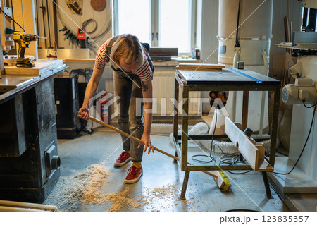 Carpenter woman cleanup workshop after woodwork. Handywoman sweep sawdust with broom, organize space Carpenter woman cleanup workshop after woodwork. Handywoman sweep sawdust with broom, organize space 123835357