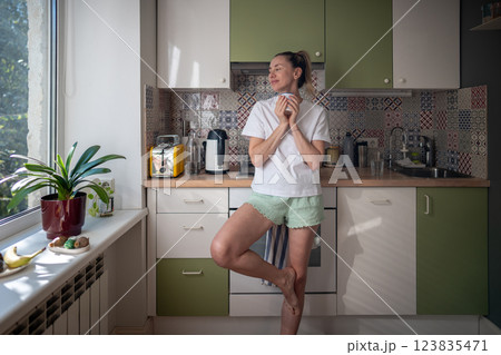 Peaceful woman standing in kitchen, holding cup of hot beverage and dreamy looks out of window. 123835471