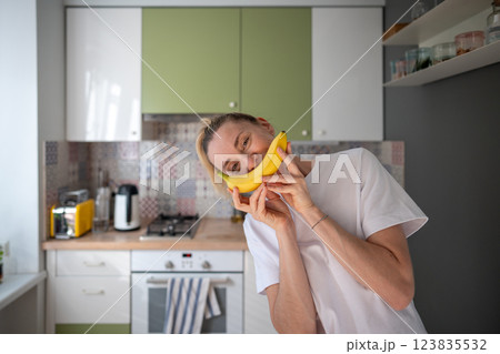 Funny goofy woman puts banana to face like smile in home kitchen. Organic healthy diet, morning meal Funny goofy woman puts banana to face like smile in home kitchen. Organic healthy diet, morning meal 123835532