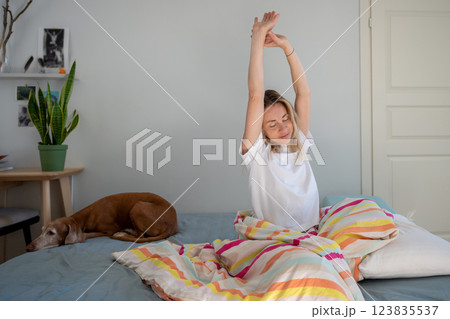 Rested woman stretching in bed while dog lounges next to her, enjoying slow mindful morning 123835537