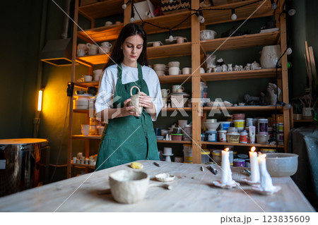 Female potter check molded clay mug for strength, touching handle in workshop. Craftsmanship skills 123835609