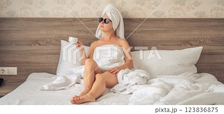 Young beautiful woman with a towel on her head sitting on the bed with a cup of coffee. 123836875