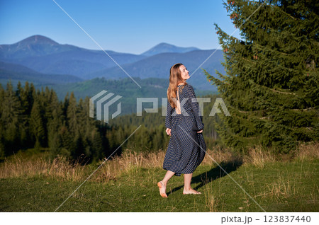 Woman in backless polka dot dress walks barefoot through meadow, holding her dress as she twirls. She enjoying stunning view of lush green forests and distant mountains under clear blue sky. Woman in backless polka dot dress walks barefoot through meadow, holding her dress as she twirls. She enjoying stunning view of lush green forests and distant mountains under clear blue sky. 123837440