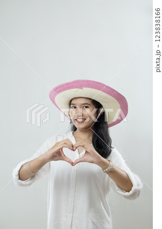 woman forming a heart towards the camera in white shirt and straw hat, woman's affectionate shape, white isolated 123838166