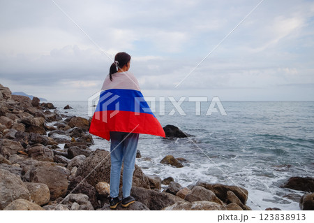A brunette woman with a Russian flag on her shoulders stands on a rocky seashore. Independence day, patriotic holiday concept A brunette woman with a Russian flag on her shoulders stands on a rocky seashore. Independence day, patriotic holiday concept 123838913