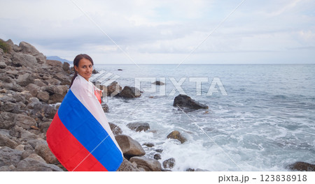 A brunette woman with a Russian flag on her shoulders stands on a rocky seashore. Independence day, patriotic holiday concept A brunette woman with a Russian flag on her shoulders stands on a rocky seashore. Independence day, patriotic holiday concept 123838918