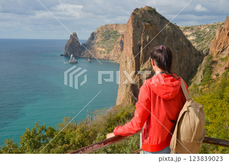 A tourist with a backpack travels through the Crimean village of Novy Svet, Cape Fiolent. Girl on the edge of a cliff enjoying the sea view. Active summer holiday, adventure lifestyle. A tourist with a backpack travels through the Crimean village of Novy Svet, Cape Fiolent. Girl on the edge of a cliff enjoying the sea view. Active summer holiday, adventure lifestyle. 123839023