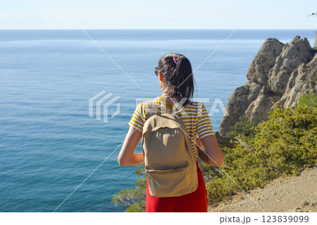Happy female tourist with a backpack on the top of a mountain, enjoying the sea view, standing on a rock and looking into the distance. The village of Novy Svet in Crimea. Russia Happy female tourist with a backpack on the top of a mountain, enjoying the sea view, standing on a rock and looking into the distance. The village of Novy Svet in Crimea. Russia 123839099