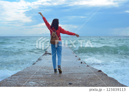 Rear view of a young woman tourist with a backpack standing on the pier and waves hitting the pier in a storm. City of Sudak, Crimea. 123839113