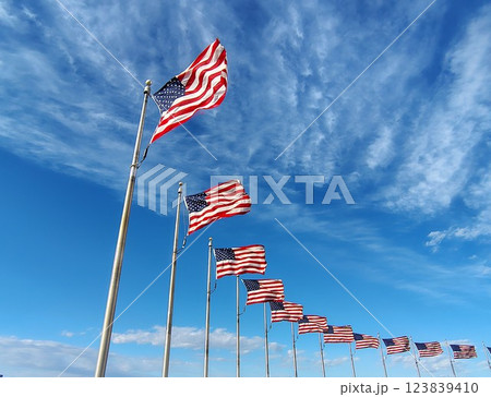 American flags on long flagpoles fluttering in the wind against a bright blue sky, national banners of the USA surrounding a monument, a symbol of the nation and state of the US 123839410