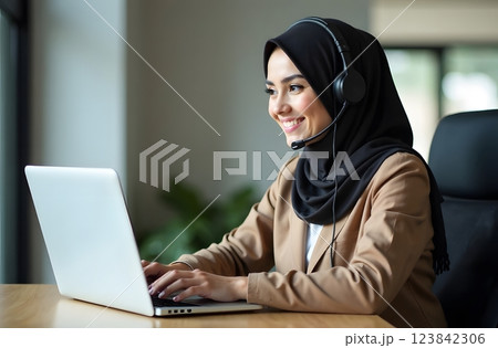 A smiling woman in a hijab wearing a headset sits at a computer desk in a bright office environment. A smiling woman in a hijab wearing a headset sits at a computer desk in a bright office environment. 123842306