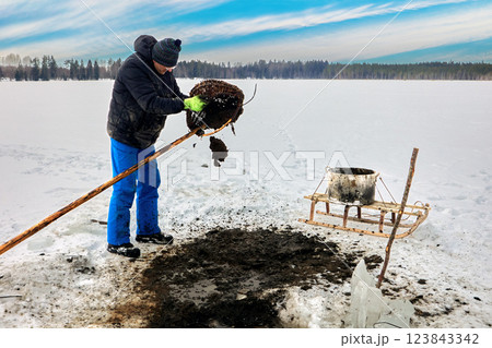 Farmer extracts lake silt sapropel using basket on pole via ice hole for agriculture, transferring organic material to sled, enriching soil naturally. 123843342