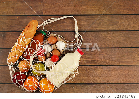 Fabric string bag with various natural products. Milk, eggs, baguettes, natural juice and fruits in a cotton bag on a wooden background, food delivery concept, advertising for a store. Fabric string bag with various natural products. Milk, eggs, baguettes, natural juice and fruits in a cotton bag on a wooden background, food delivery concept, advertising for a store. 123843468