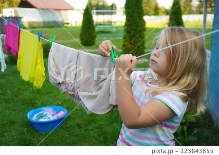 Wearing a cheerful summer dress, a six-year-old girl stretches her small arms to hang a damp shirt, her face full of focus. 123843655