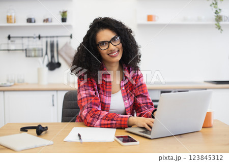 Smiling African woman with curly hair, wearing red shirt, working on laptop, at home kitchen. Scene conveys comfort, productivity, and warmth. Smiling African woman with curly hair, wearing red shirt, working on laptop, at home kitchen. Scene conveys comfort, productivity, and warmth. 123845312
