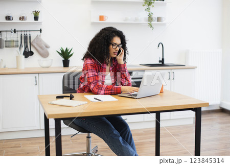 Young African woman wearing casual clothes using laptop and phone, working from kitchen setting 123845314