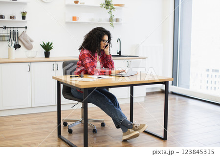 African woman sitting at desk in bright kitchen using technology, appearing focused and professional. Scene represents remote work, productivity, and comfortable home office. 123845315
