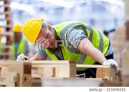 Wood factory construction worker wearing a yellow hard hat and safety vest focuses on measuring and crafting wooden materials in well-lit workshop. Wood factory construction worker wearing a yellow hard hat and safety vest focuses on measuring and crafting wooden materials in well-lit workshop. 123846581