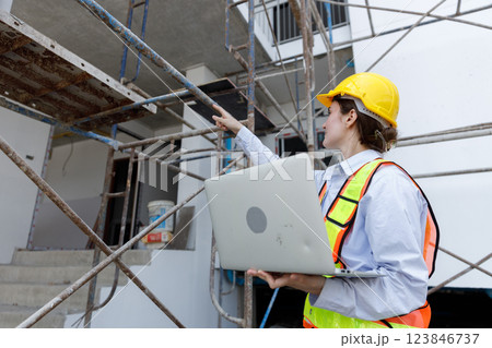 Construction worker stands confidently at a building site, wearing a bright yellow helmet and safety vest while using a laptop to monitor project progress during day. Construction worker stands confidently at a building site, wearing a bright yellow helmet and safety vest while using a laptop to monitor project progress during day. 123846737