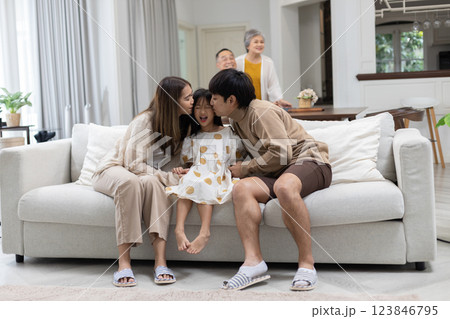 happy family shares a loving moment in their living room. Parents kiss their daughter, who beams with joy, while a grandparent looks on fondly, creating a warm atmosphere. 123846795