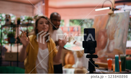 Woman artist hosting online class to present products and technique used on rustic artwork, recording tutorial online. Team of painters doing master class for e learning, art instruction. Camera B. 123848898