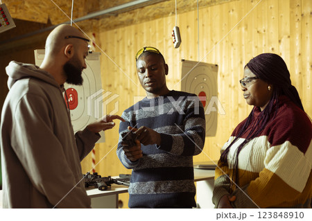 Shooting gallery employee teaching students how to cock pistol before firing. African american firing range instructor providing guide to achieve high level of proficiency with weapons Shooting gallery employee teaching students how to cock pistol before firing. African american firing range instructor providing guide to achieve high level of proficiency with weapons 123848910