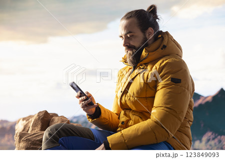 Traveler in warm clothing scrolling social media on top of the mountain peak, person always connected with a strong signal and obsessed with technology. Climbing the rocky hills. 123849093