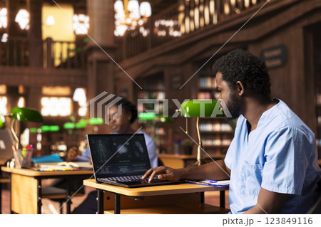 Black med school scholar studying and taking notes using online information, reviews a thesis project in the campus library. Young adult collecting academic data for medical research. 123849116
