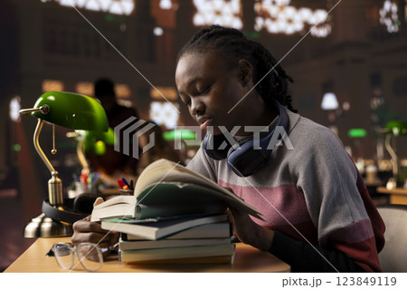 African american sleepy girl studying with exhaustion and burnout all night for exams, making preparations for a difficult school subjects. Young student consulting specialty books in the library. 123849119