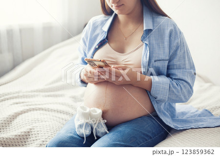 A pregnant woman is seen resting comfortably on bed, holding a pair of baby shoes near her belly. She appears content and serene, enjoying a quiet moment 123859362