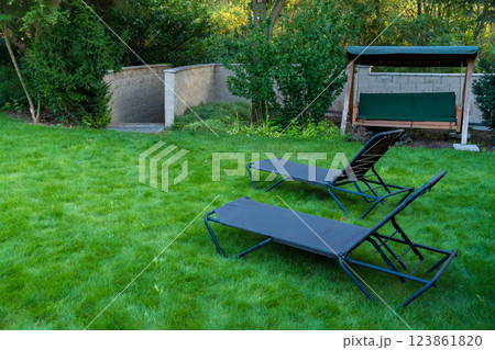 Peaceful backyard featuring two garden loungers and a swing bench set on a lush green lawn Peaceful backyard featuring two garden loungers and a swing bench set on a lush green lawn 123861820