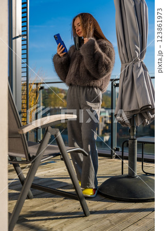 Smiling woman on a sunny terrace in autumn Prague, holding coffee and smartphone in a faux fur coat 123861973
