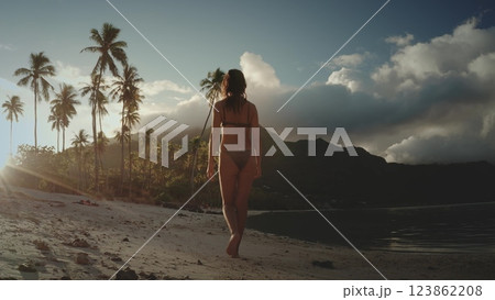 A woman walks barefoot on a white sand beach in French Polynesia, with a line of palm trees and a mountain in the background. The sun is setting, casting a golden glow on the scene. 123862208