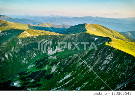 Aerial view of rugged mountain range with lush green slopes at sunset. Landscape showcases beauty of untouched nature with distant peaks and valleys bathed in soft sunlight. Carpathians, Chornogora. 123863545
