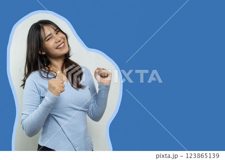 A girl stands smiling happily celebrating her success. She is wearing a casual outfit and has long wavy hair, isolated on plain blue. 123865139