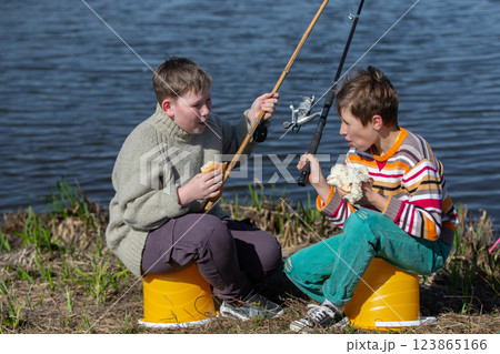 The boys are friends on a fishing trip by the river eating a bun and laughing. Children are friends on a summer fishing trip. 123865166