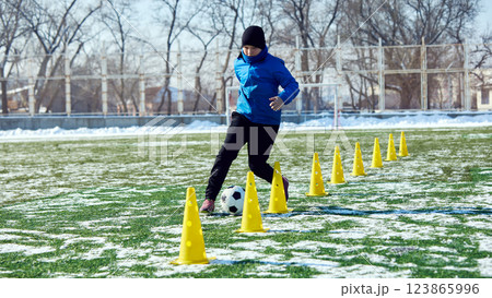 Football player dribbling soccer ball through yellow cones on outdoor field, improving control and coordination. 123865996