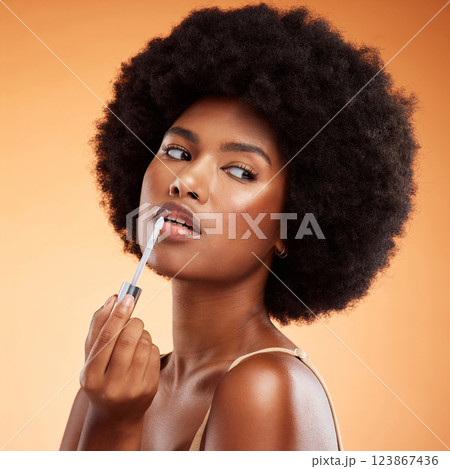 Black woman, afro and natural beauty lip gloss treatment for a healthy, shiny and transparent tint. Cosmetics, apply and beautiful face of African model holding makeup tool at orange background. Black woman, afro and natural beauty lip gloss treatment for a healthy, shiny and transparent tint. Cosmetics, apply and beautiful face of African model holding makeup tool at orange background. 123867436