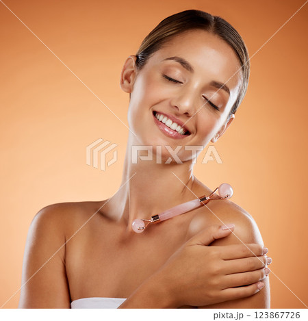 Cosmetics, woman and face roller for natural beauty, skincare and smile with brown studio background. Makeup, body care and confident girl being happy, with smooth skin and dermatology wellness. 123867726