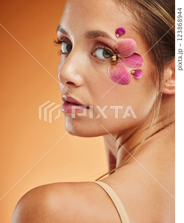 Model, eye and flowers with makeup in studio portrait for fashion, cosmetics or beauty against backdrop. Woman, art and flower with petals on skin for skincare, happy and face orange background 123868944