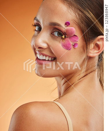 Beauty, makeup and creative with woman petals on face for design, flowers and natural cosmetics against orange background in studio. Spring, skincare and smile with girl for color, model and orchid 123868947