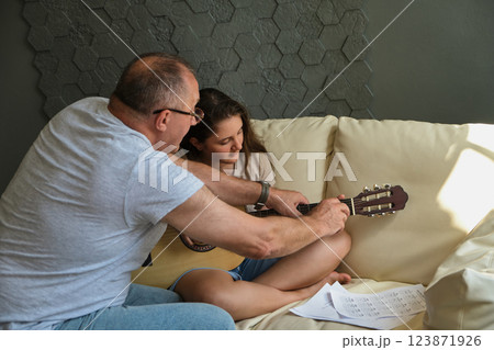 Grandpa teaching his granddaughter to play guitar on sofa at home 123871926