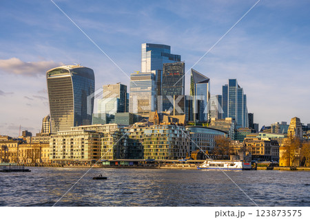 Modern skyscrapers rise against a clear sky in London City, reflecting on the River Thames. Boats glide along the water, showcasing the vibrant urban landscape of England. 123873575