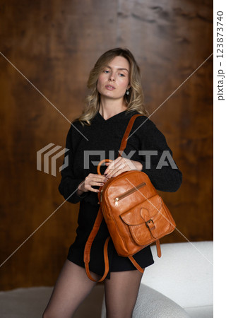 A girl in a warm sweater, a black short skirt and nylon tights is posing with a brown backpack. A girl in a warm sweater, a black short skirt and nylon tights is posing with a brown backpack. 123873740