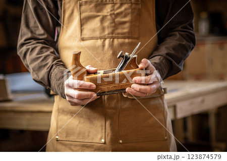 Carpenter wearing brown apron holding vintage wooden hand plane in workshop Carpenter wearing brown apron holding vintage wooden hand plane in workshop 123874579
