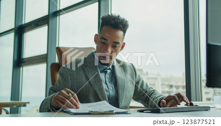 A focused businessman taking notes at his desk in a modern office with large windows, highlighting professionalism and attention to detail. 123877521