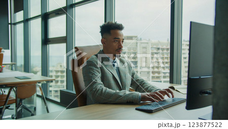 A focused businessman working on a computer in a modern office with floor-to-ceiling windows overlooking an urban cityscape, representing professionalism, technology, and workplace efficiency. 123877522
