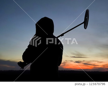 The silhouette of a hooded treasure hunter at sunset. Treasure Hunter - Gold digger - Man Holding Metal Detector The silhouette of a hooded treasure hunter at sunset. Treasure Hunter - Gold digger - Man Holding Metal Detector 123878796